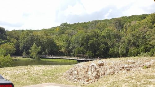 A scenic view of a bridge over a calm river, surrounded by lush green trees and a grassy area.