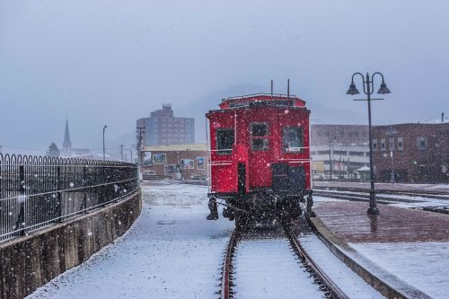 A red train car on snowy tracks, surrounded by a winter landscape and buildings in the background. Snowflakes are falling.