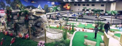 Indoor amusement park scene featuring a waterfall, mini-golf, and go-kart track surrounded by tropical decor.