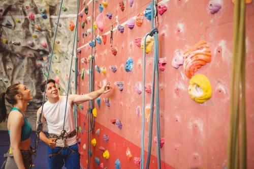 Two climbers at an indoor rock climbing wall, discussing routes while harnessed and preparing to climb.