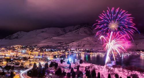 Colorful fireworks illuminate a snowy landscape and a lakeside town at night, with mountains in the background.