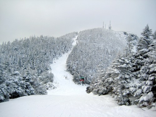 Snow-covered mountain with evergreen trees and a ski slope, under a cloudy sky. Ski lift visible in the distance.
