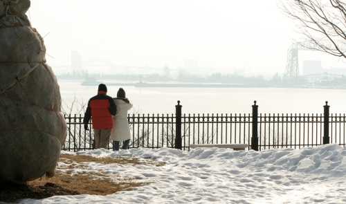 A couple stands by a fence overlooking a foggy river, with snow on the ground and a large sack nearby.