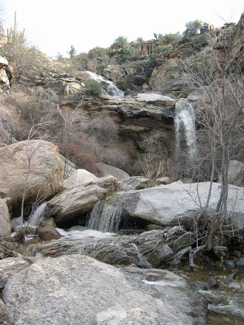 A serene waterfall cascades over rocky cliffs, surrounded by dry vegetation and boulders in a natural landscape.