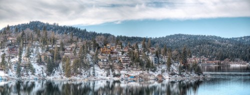 A snowy lakeside village with houses nestled among trees, reflecting on the calm water under a cloudy sky.