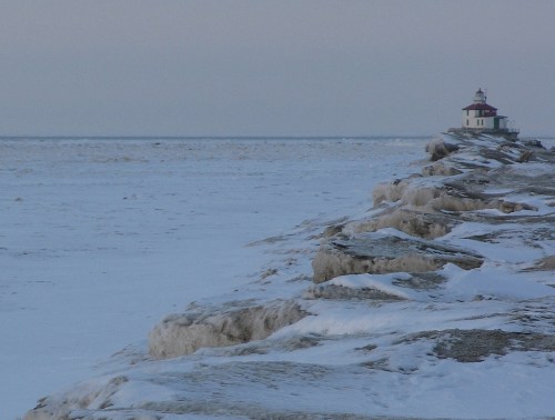 A snowy pier extends into a frozen lake, leading to a distant lighthouse under a pale sky.