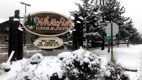 Sign for Whitefish Lodge & Suites, surrounded by snow-covered trees, with a street sign nearby.