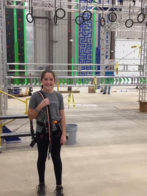 A young girl stands in an indoor obstacle course, wearing a harness and smiling, with climbing rings in the background.