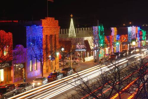 Colorful holiday lights adorn buildings along a street at night, with a Christmas tree illuminated in the background.