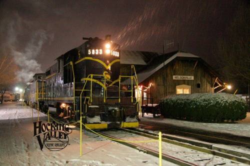A snow-covered train station at night, with a vintage locomotive parked outside and snow gently falling.
