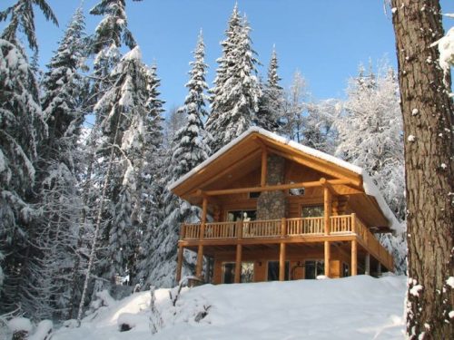 A cozy wooden cabin surrounded by snow-covered trees under a clear blue sky.