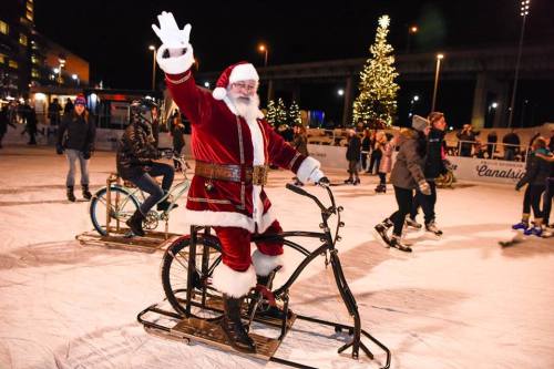 Santa Claus waves while riding a bike on ice, surrounded by skaters and festive holiday decorations.
