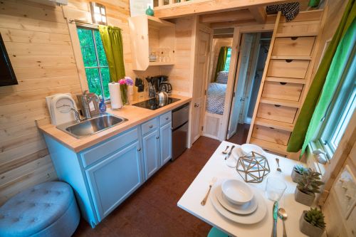 Cozy tiny home interior featuring a kitchen, dining area, and ladder to a loft, with wooden walls and green accents.