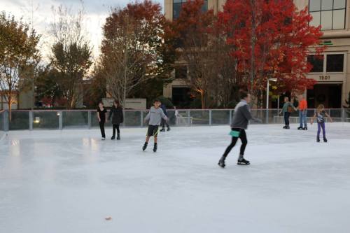A group of people ice skating on an outdoor rink surrounded by autumn trees and buildings.