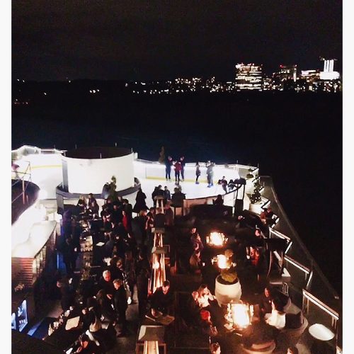 A rooftop bar scene at night, filled with people socializing, with city lights in the background.