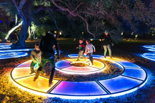 Children and adults play on illuminated circular platforms in a park at night, surrounded by trees and colorful lights.