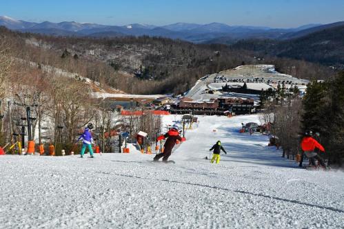 Skiers descend a snowy slope with a mountain backdrop and a ski resort in the distance on a clear day.