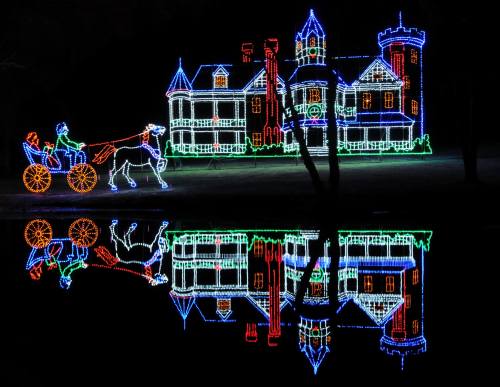 Colorful holiday lights illuminate a castle and a horse-drawn carriage, reflecting in a calm water surface.