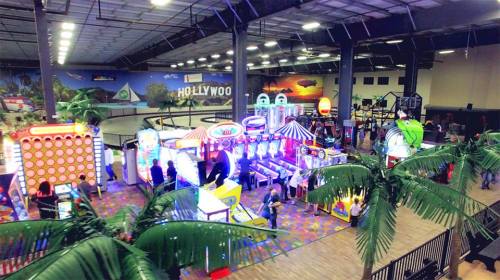A vibrant indoor arcade with colorful games, bright lights, and palm trees, bustling with people enjoying various attractions.