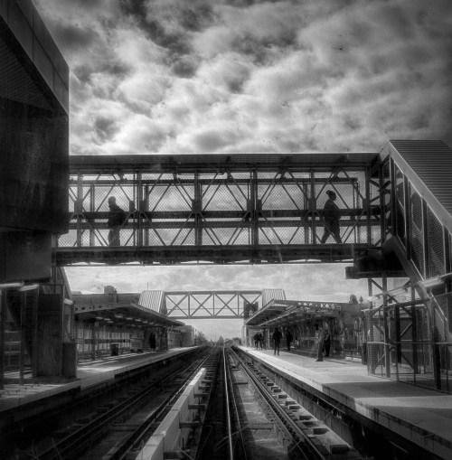 A black and white image of a train station with a pedestrian bridge and tracks extending into the distance under a cloudy sky.