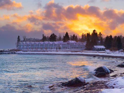 A serene winter sunset over a snowy beach, with a cozy hotel and trees in the background. Waves gently lap at the shore.