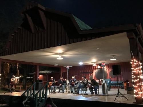 A band performs on an outdoor stage at night, surrounded by festive lights and a rustic building backdrop.