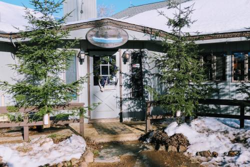 A cozy building with a sign, surrounded by snow and evergreen trees, featuring a welcoming entrance.