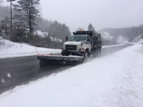 A snowplow clears a snowy road during a winter storm, with trees and hills in the background.