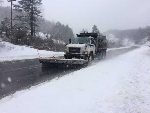 A snowplow clears a snowy road during a winter storm, with trees and hills in the background.