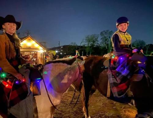 A woman and a child on decorated horses, wearing festive lights, at a nighttime event with a cozy cabin in the background.