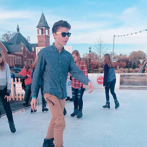 A group of people ice skating outdoors, with a young man in sunglasses and a button-up shirt in the foreground.