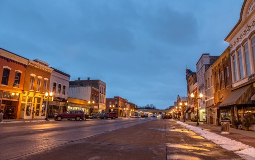 A quiet, snow-covered street at dusk, lined with historic buildings and warm streetlights illuminating the scene.