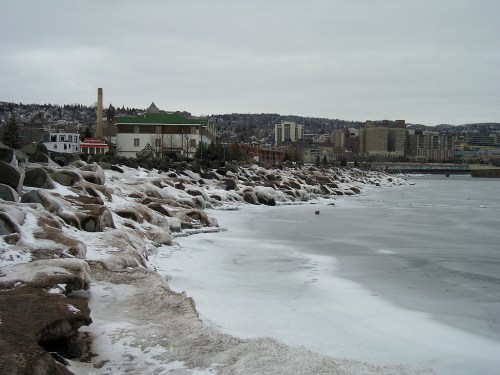 A rocky shoreline with ice and snow, overlooking a city skyline under a cloudy sky.