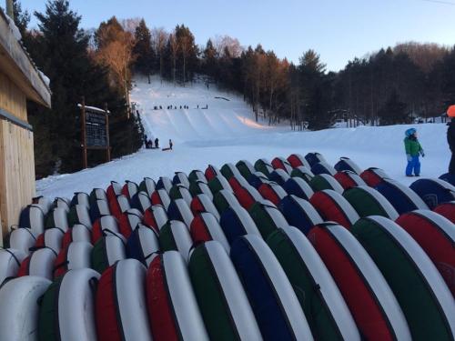 A stack of colorful snow tubes in front of a snowy hill with people sledding in the background.