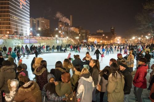 A bustling outdoor ice skating rink at night, filled with skaters and spectators in a city setting.