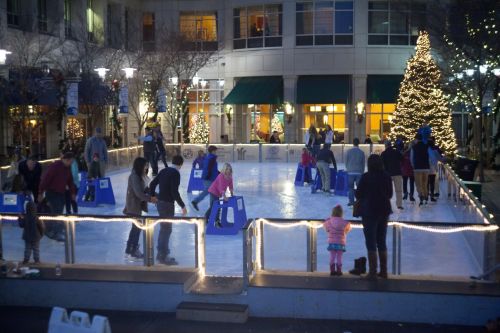 People ice skating at a festive outdoor rink, with a decorated tree and lights in the background.