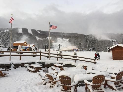 Snow-covered ski resort with American flags, wooden chairs, and ski slopes in the background.
