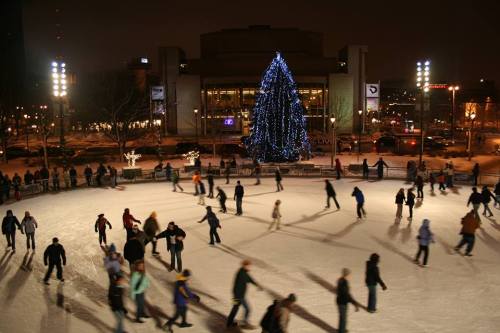 A bustling ice skating rink at night, decorated with a large blue-lit Christmas tree in the background.