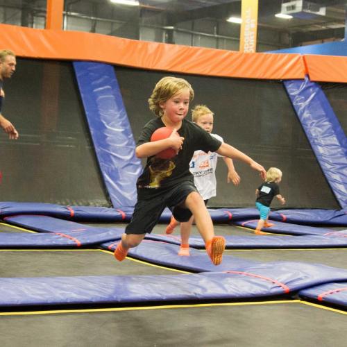 A young boy jumps on a trampoline while holding a red ball, with other children playing in the background.