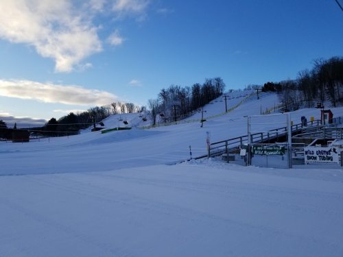 Snow-covered ski slope with a clear blue sky, ski lifts in the background, and a wooden railing in the foreground.