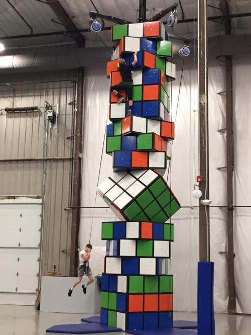 Two people climbing a large, colorful Rubik's Cube structure indoors, with one person jumping down.