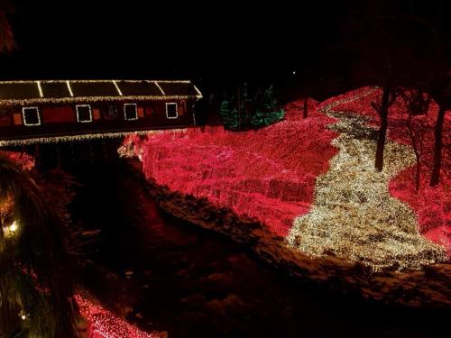 A beautifully lit scene at night featuring a covered bridge and a riverbank adorned with colorful holiday lights.