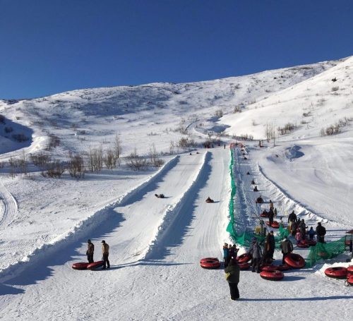 A snowy landscape with people tubing down slopes and a conveyor belt for easy access to the top. Clear blue sky above.