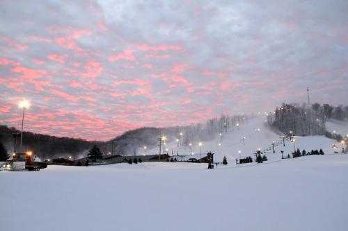 A snowy landscape at dusk, with pink clouds and ski slopes illuminated by lights in the background.