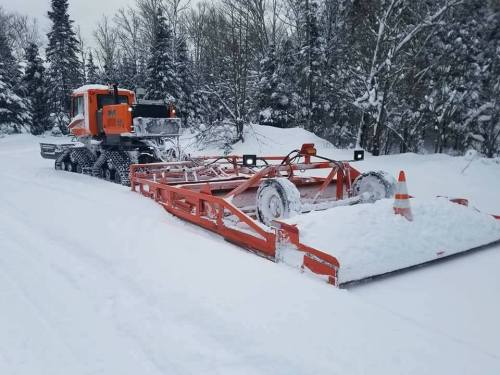 A snowplow truck clearing a snowy road surrounded by trees in a winter landscape.