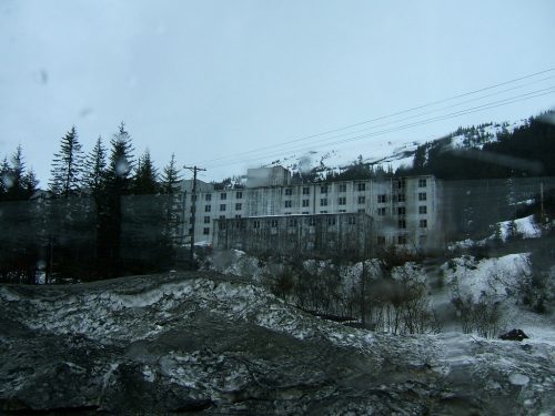 Abandoned building surrounded by snow-covered mountains and trees, with a cloudy sky overhead.
