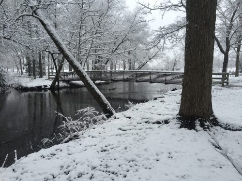 A snowy landscape featuring a wooden bridge over a calm stream, surrounded by trees and a blanket of snow.