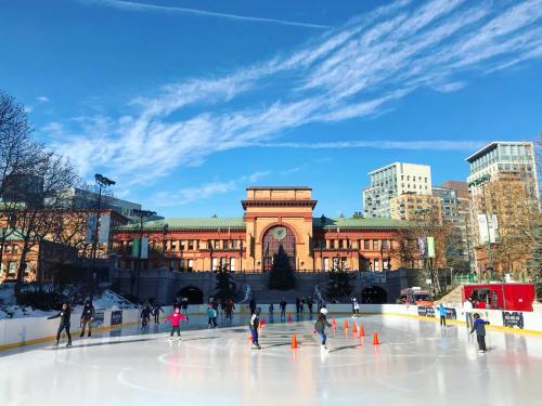 Outdoor ice skating rink with skaters, surrounded by trees and buildings under a clear blue sky.