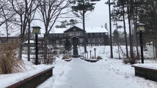 A snowy pathway leads to a large, dark wooden lodge surrounded by trees.