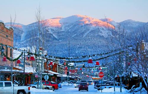 Snow-covered street in a mountain town, decorated for the holidays, with mountains in the background and a clear blue sky.